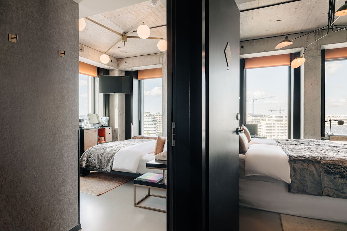 A modern hotel room with two beds, a desk, and a large window offering a city view.