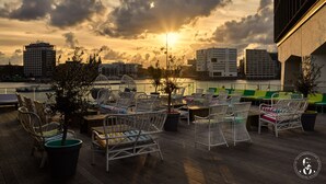 Terraza en la azotea con sillas y mesas de mimbre, con vistas a un paisaje urbano y una masa de agua al atardecer.