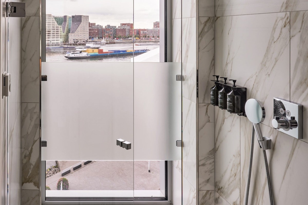 Un baño con un ventanal grande, una ducha con múltiples rociadores y vistas a un paisaje urbano.