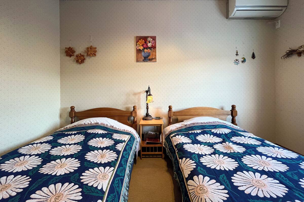 A room with two single beds, a nightstand with a lamp, a wall-mounted air conditioner, and a floral painting above the beds.