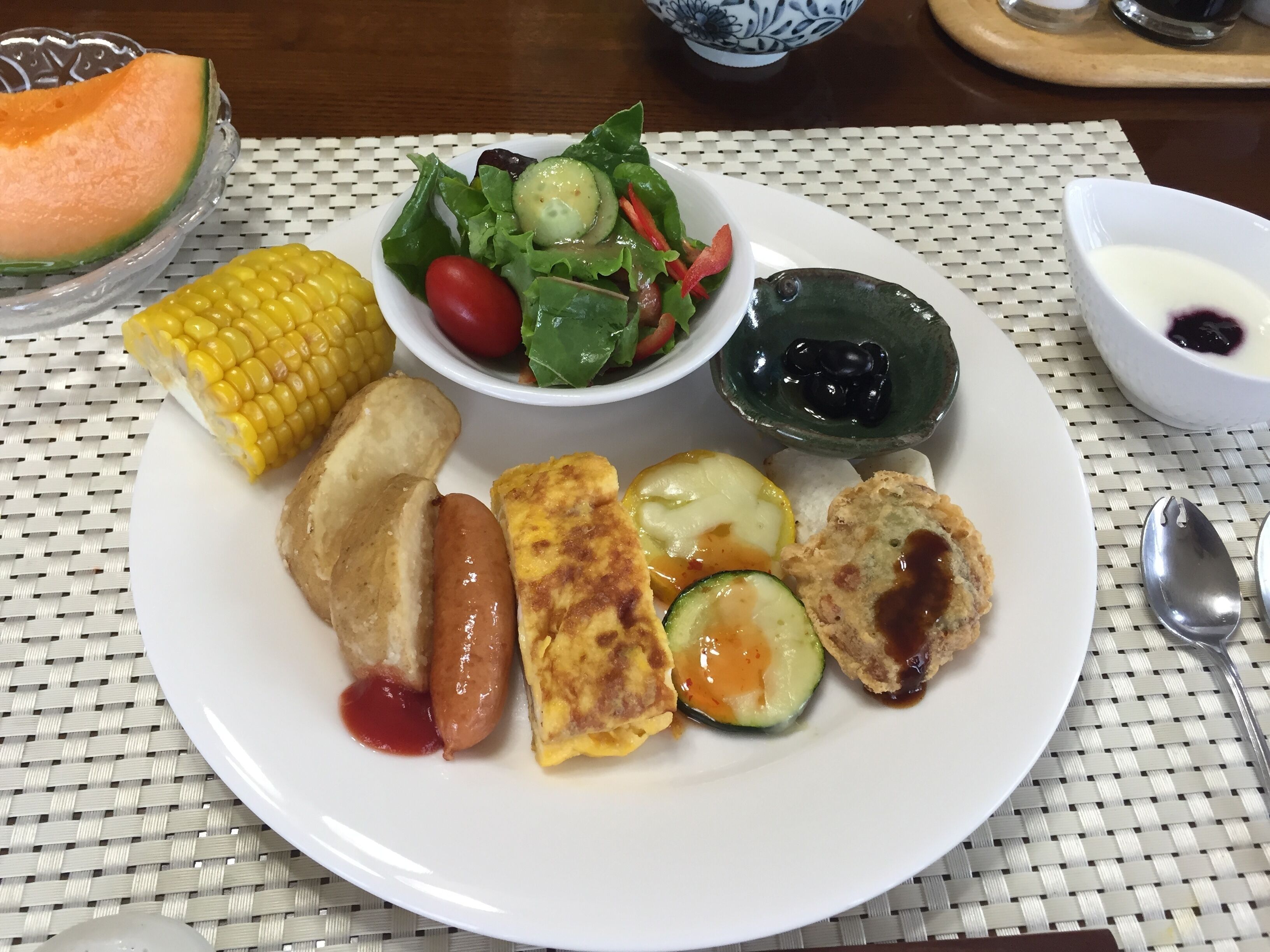 Un plato con varios alimentos, incluyendo maíz, salchicha, tortilla y ensalada.