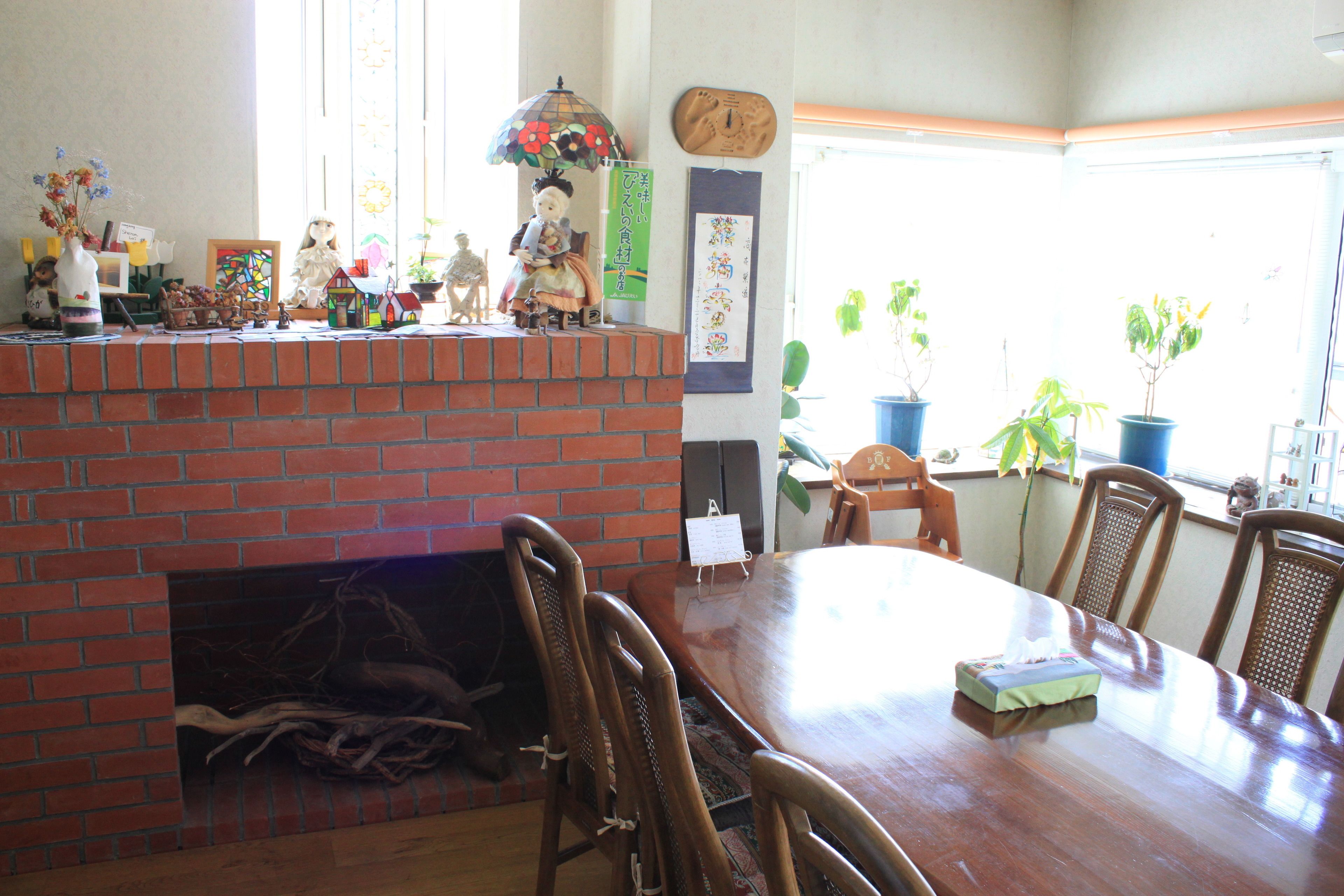 Un comedor con chimenea de ladrillo, sillas de madera, una gran mesa de madera y plantas en macetas junto a la ventana.