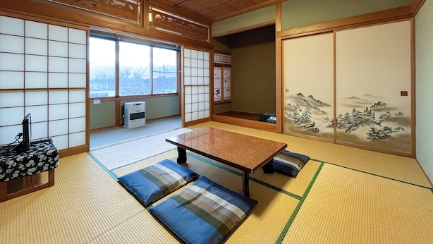 A traditional Japanese room with tatami flooring, a low table, cushions, sliding doors with artwork, and a view through a large window. - Pension Natsumi no Sato (Biei)