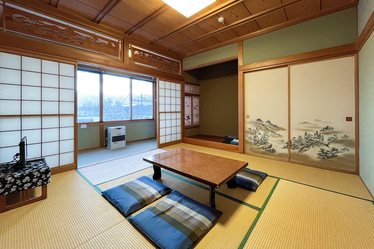 A traditional Japanese room with tatami flooring, a low table, cushions, sliding doors with artwork, and a view through a large window.