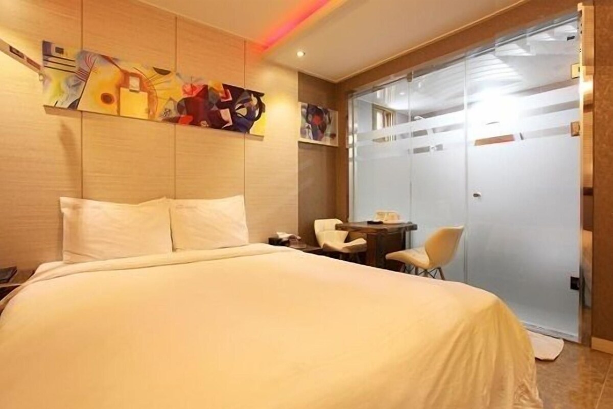 A hotel room with a large bed, a small table with chairs, and a glass partition wall.