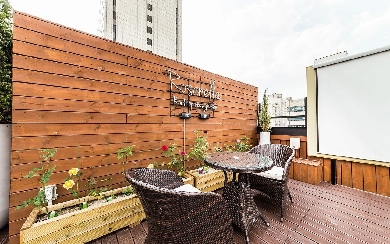 A rooftop garden with wooden decking, wicker furniture, and a sign reading "Roseholla Rooftop Rose Garden".