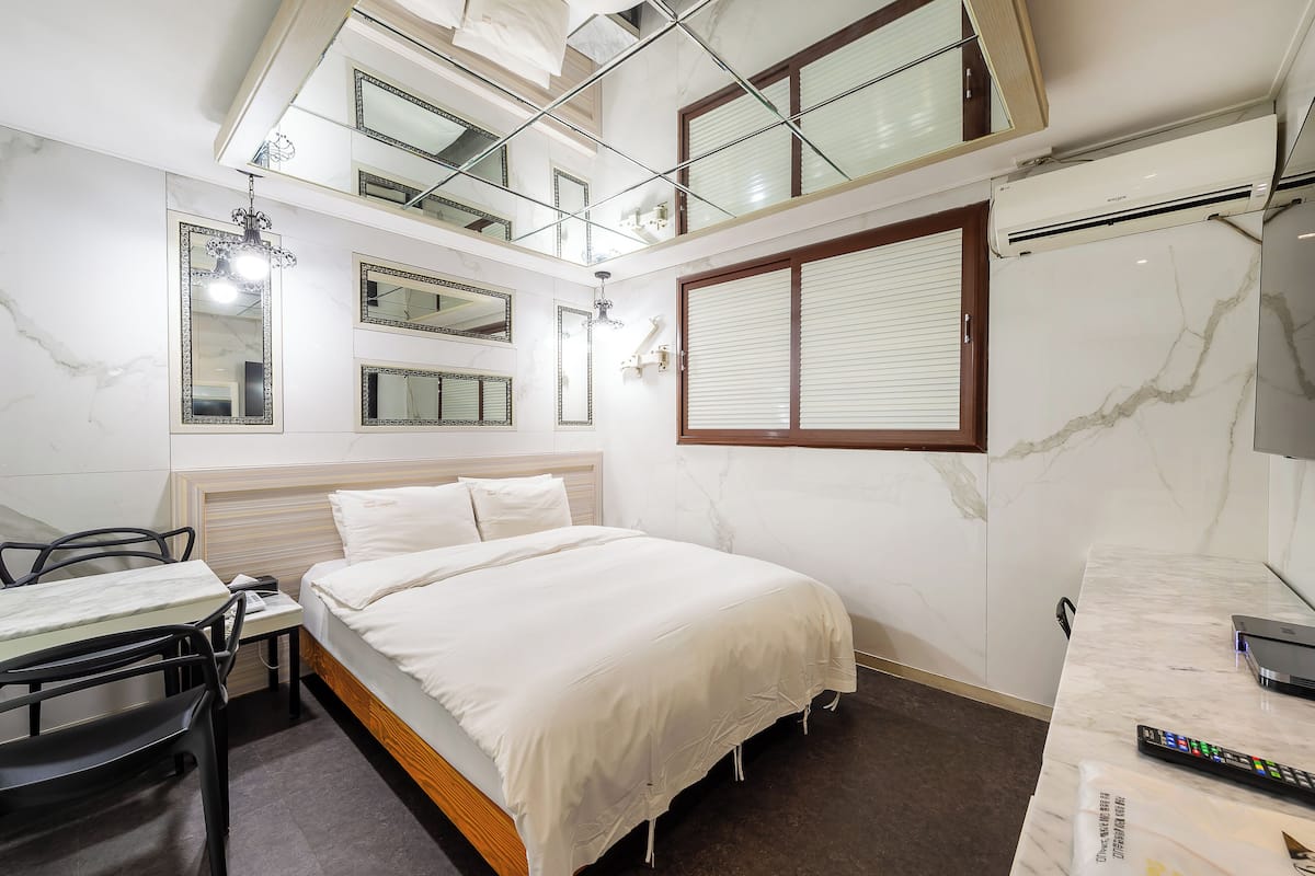 A hotel room with a bed, a desk, a chair, a mirror, a window with blinds, and an air conditioning unit.