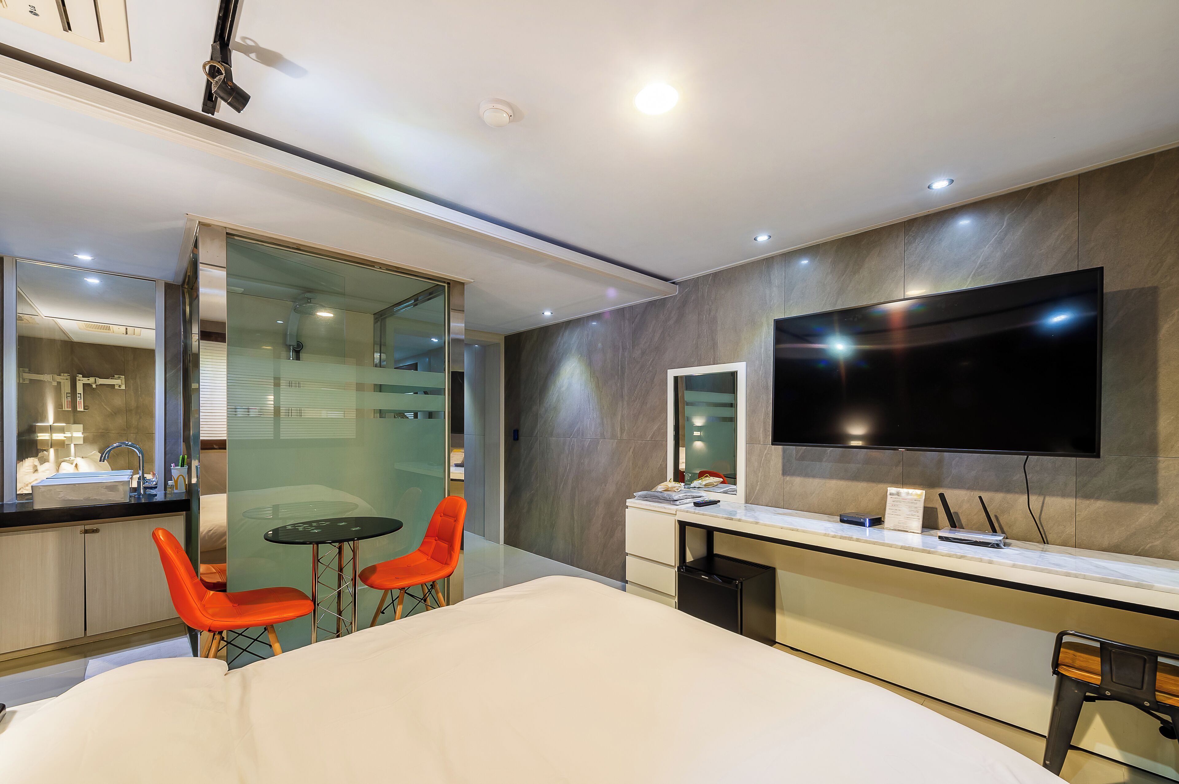A modern hotel room with a large bed, a desk with a flat-screen TV, a glass shower enclosure, and orange chairs.