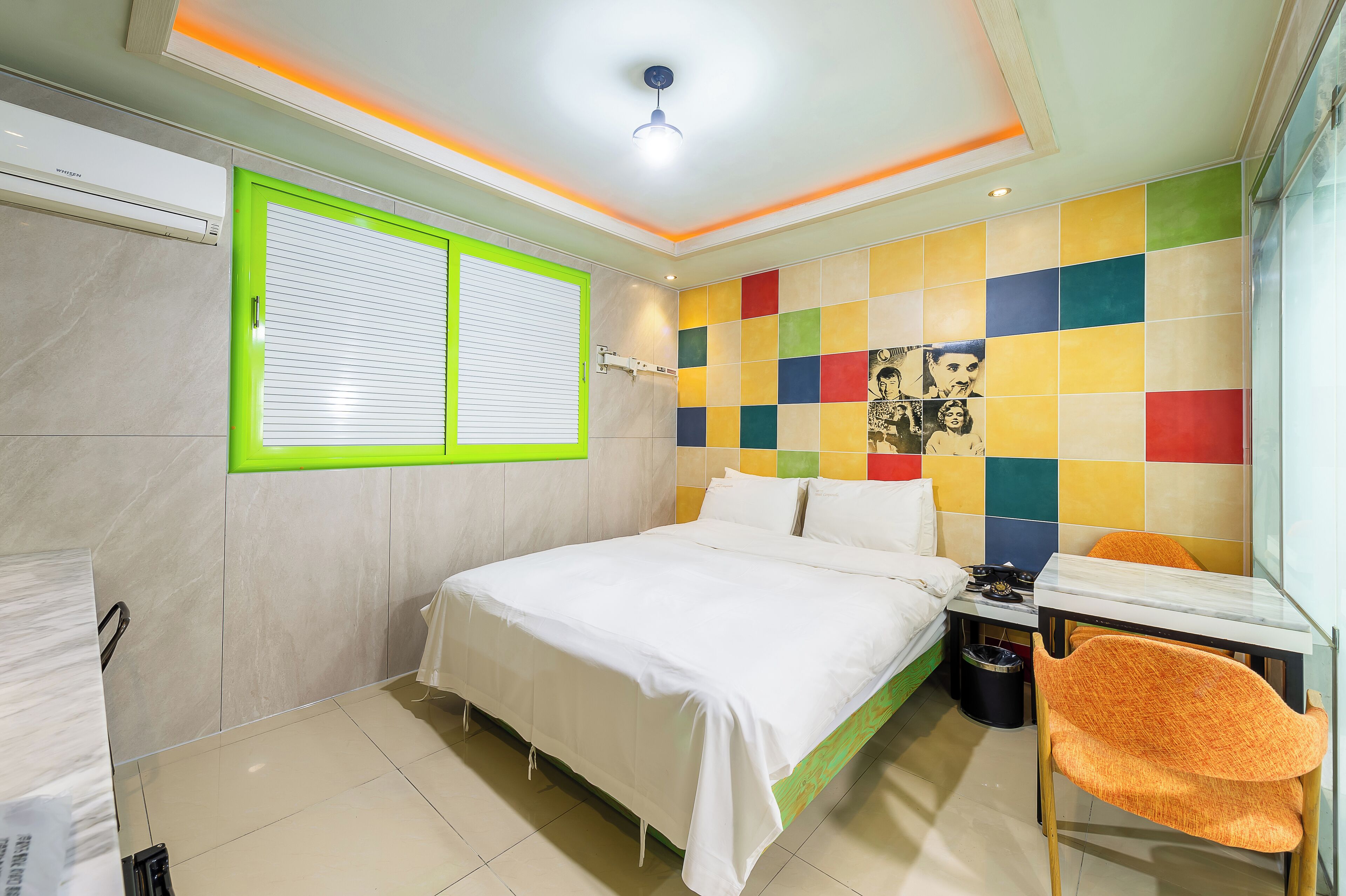 A modern bedroom with a bed, a window with blinds, a colorful wall, and a small table with a chair.