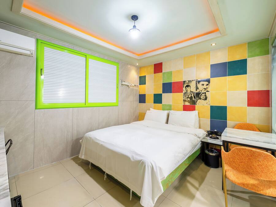 A modern bedroom with a bed, a window with blinds, a colorful wall, and a small table with a chair.