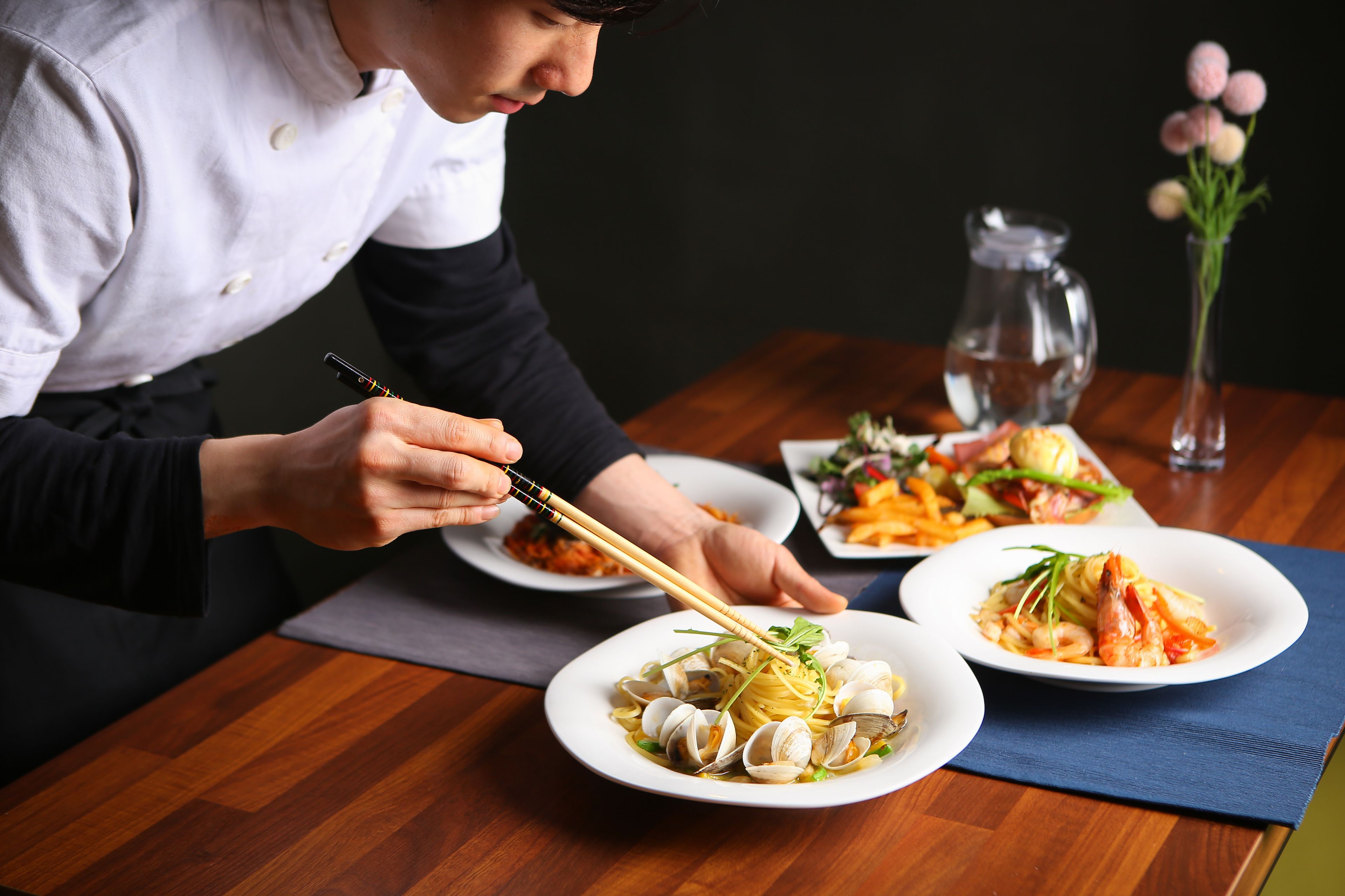 A person serving food on a wooden table with multiple dishes, including clams and shrimp, and a vase with flowers in the background.