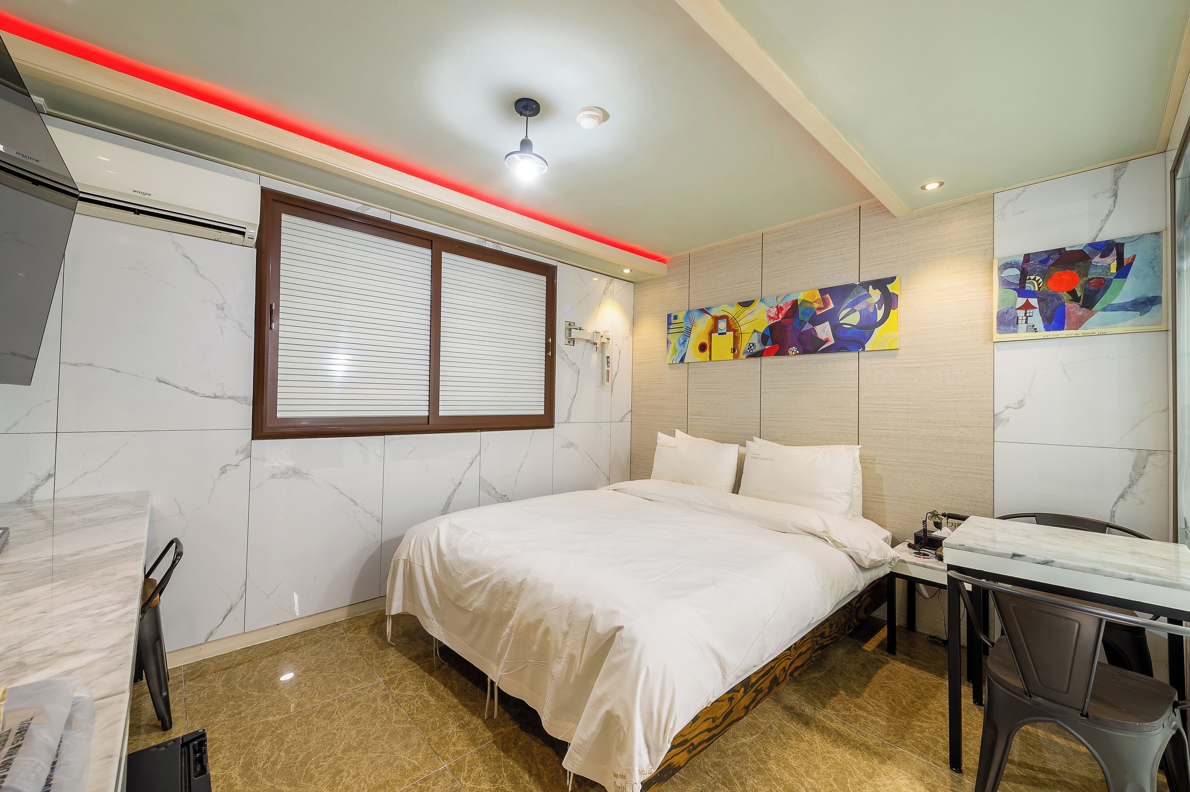 A hotel room with a bed, a desk, and a chair. There is a window with blinds, a ceiling light, and a wall-mounted air conditioning unit.