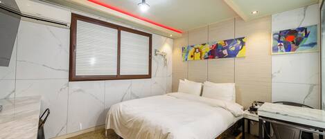 A hotel room with a bed, a desk, and a chair. There is a window with blinds, a ceiling light, and a wall-mounted air conditioning unit.