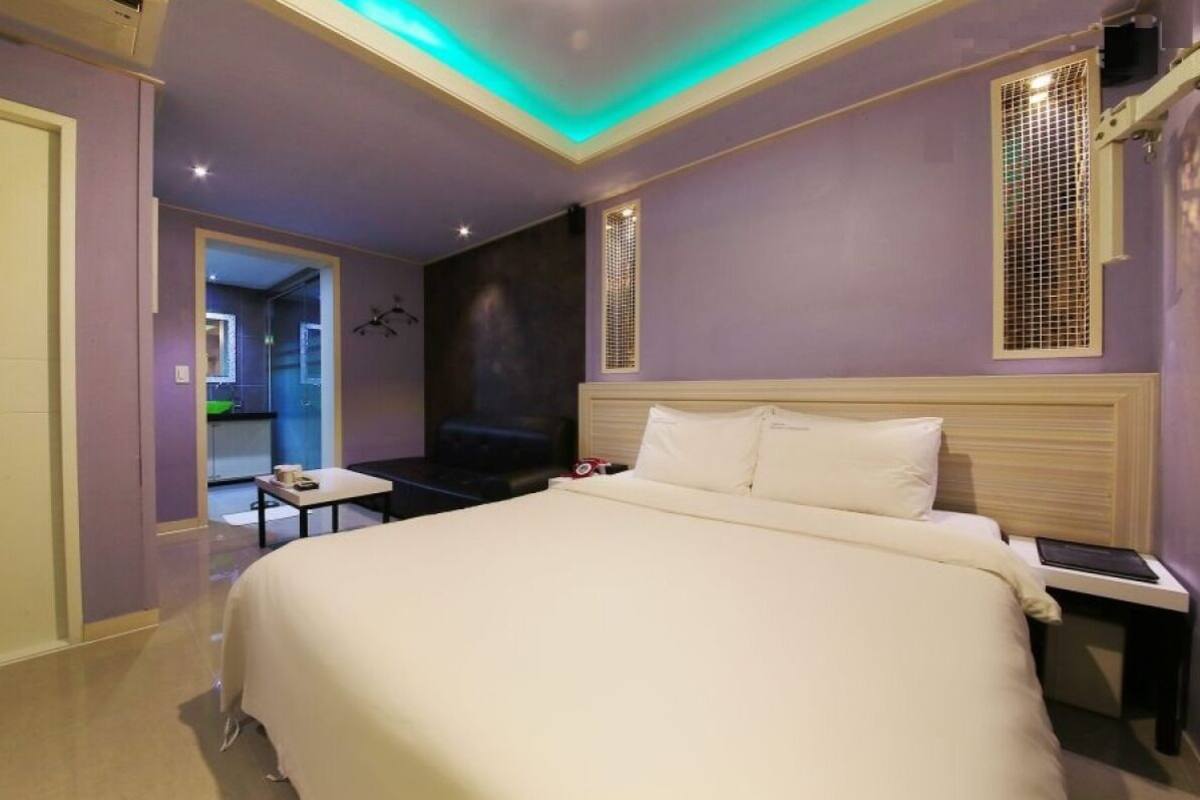 A hotel room with a large bed, a bedside table, a small table, and a chair. There is a door leading to another room and a window with a view.