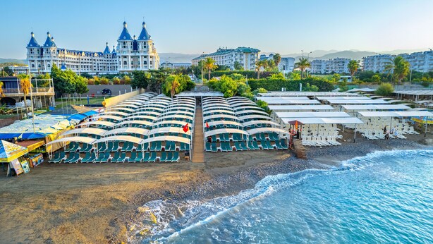 Un hotel a la vora de la platja, amb nombroses gandules i ombrel·les, un edifici gran amb teulades en forma de cúpula i vista al mar.