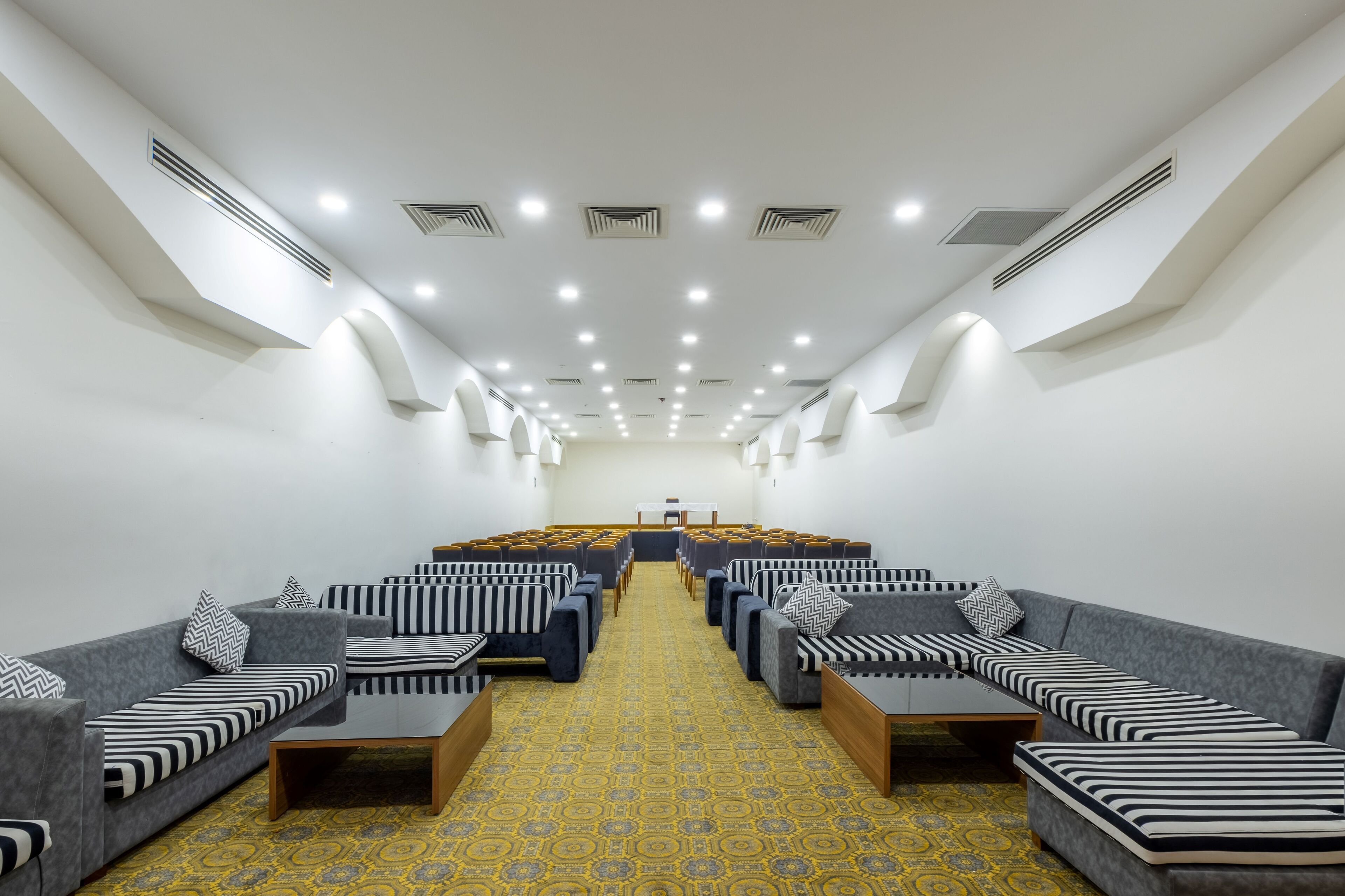 A modern hotel room with a central seating area, a patterned carpet, and a white ceiling with recessed lighting.