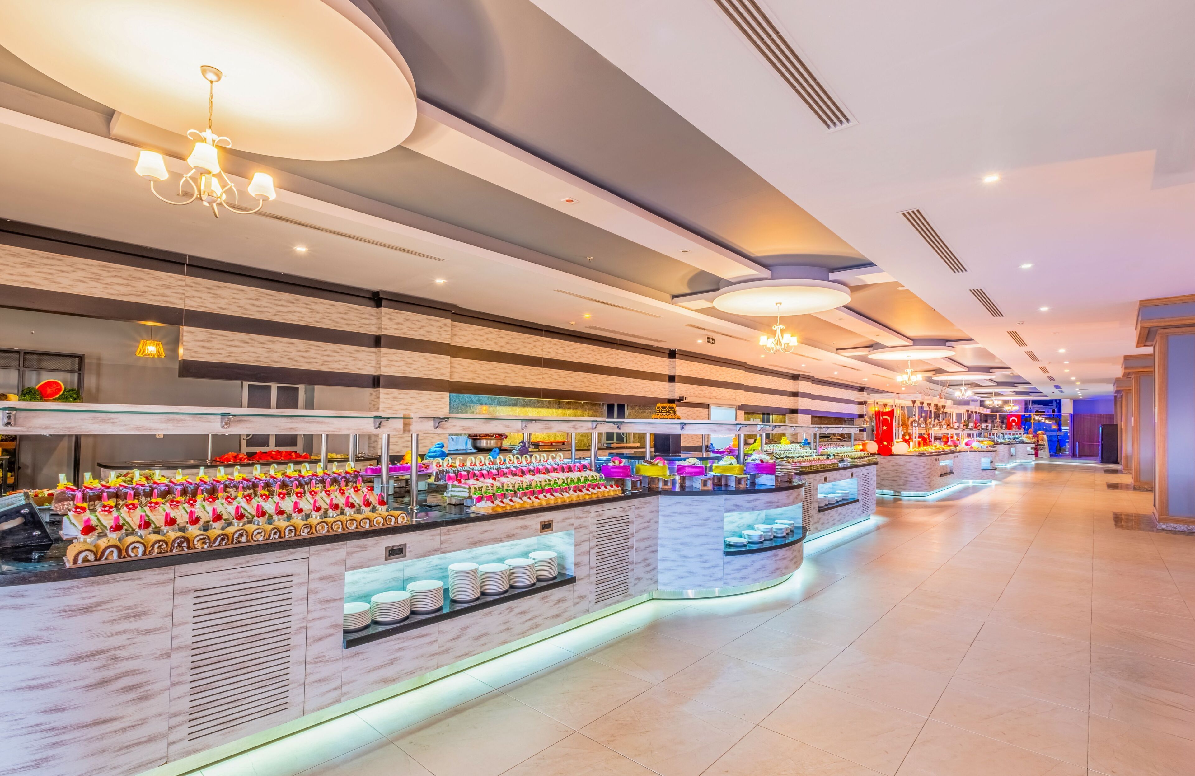 A modern hotel lobby with a long reception desk, multiple food stations, and a spacious layout.