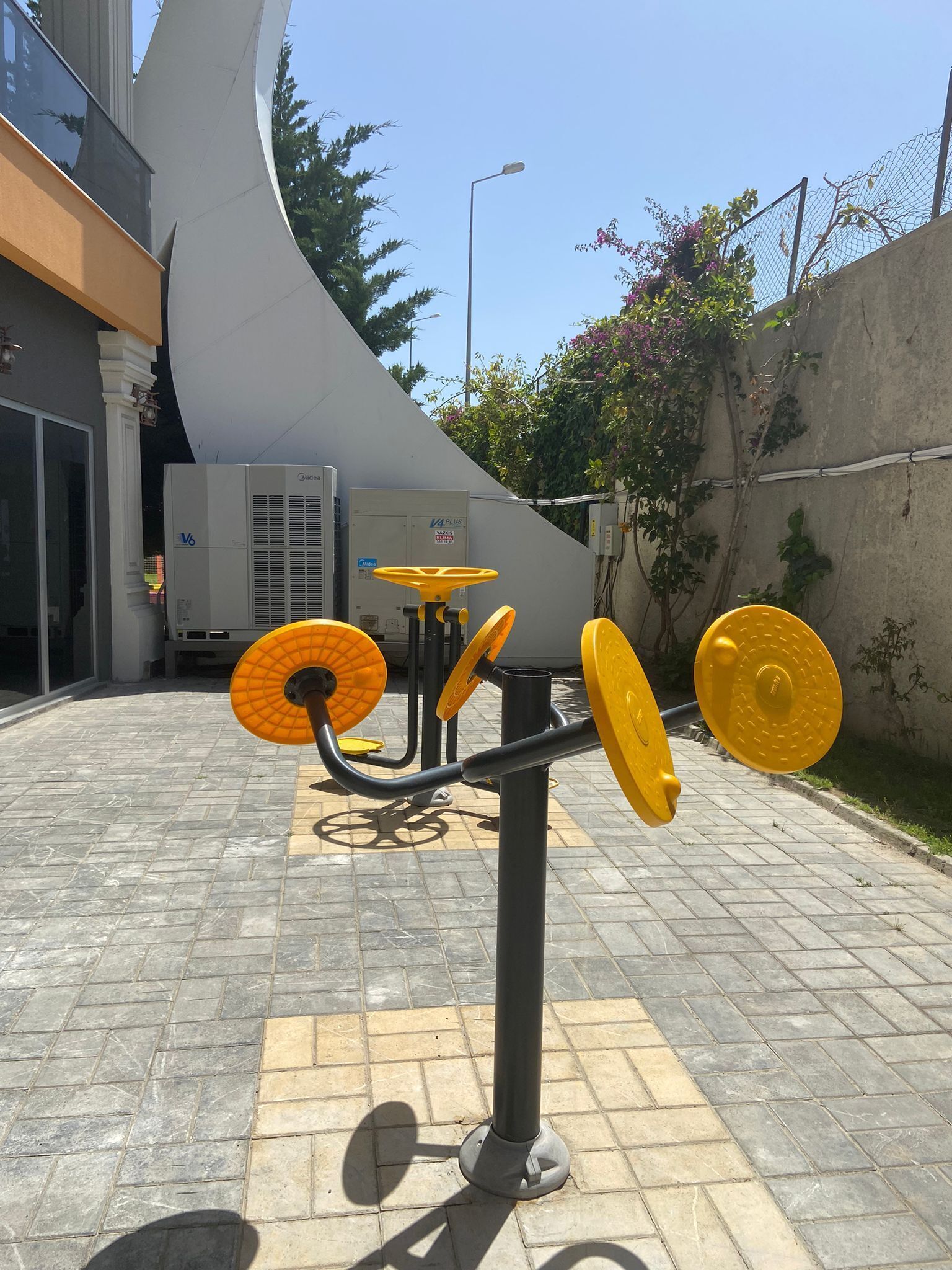 Equipamento de exercício ao ar livre com design curvo, situado em área pavimentada, com um edifício e unidades de ar condicionado ao fundo.