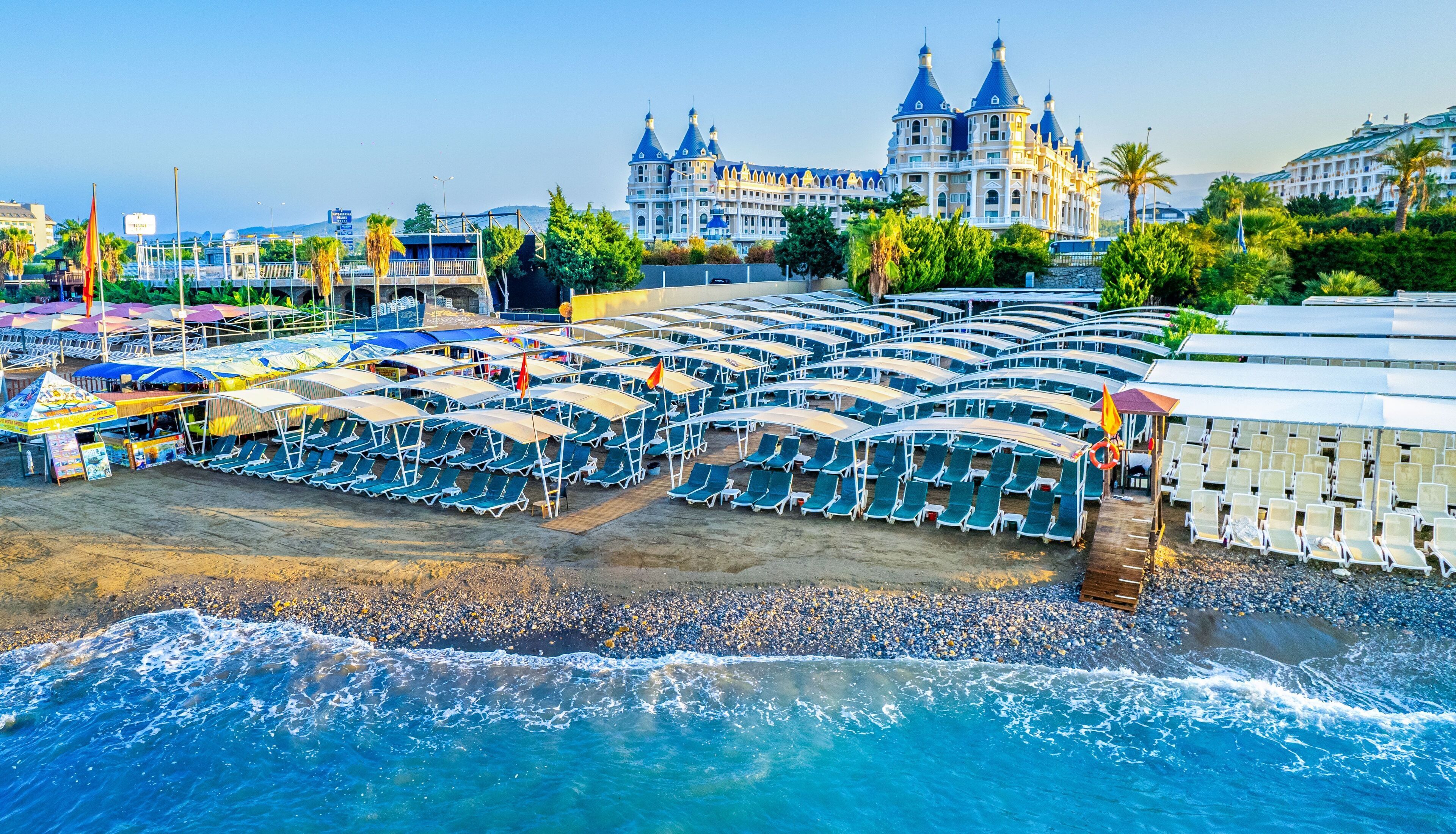 A beachside hotel with rows of sun loungers and umbrellas, a large building with domed towers, and a rocky shoreline.