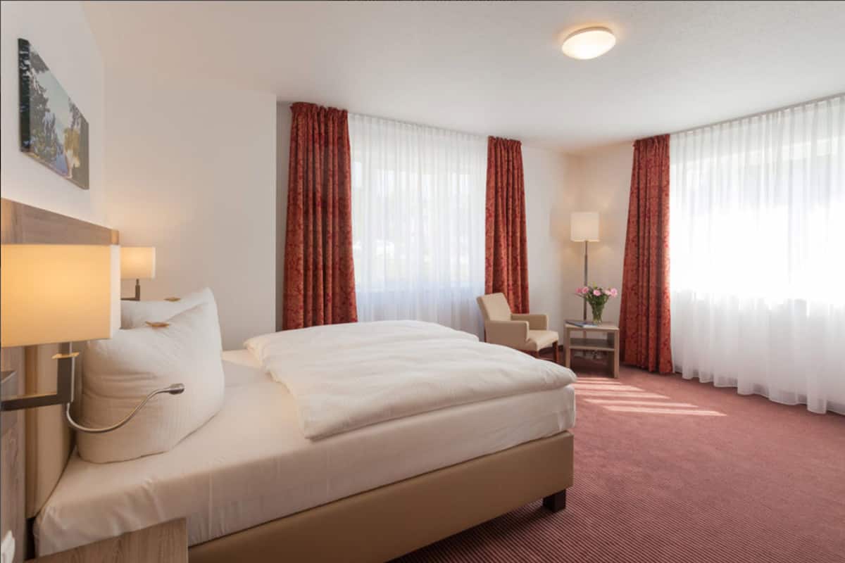 Ett hotellrum med en stor säng, ett skrivbord med en stol, en lampa och en vas med blommor.