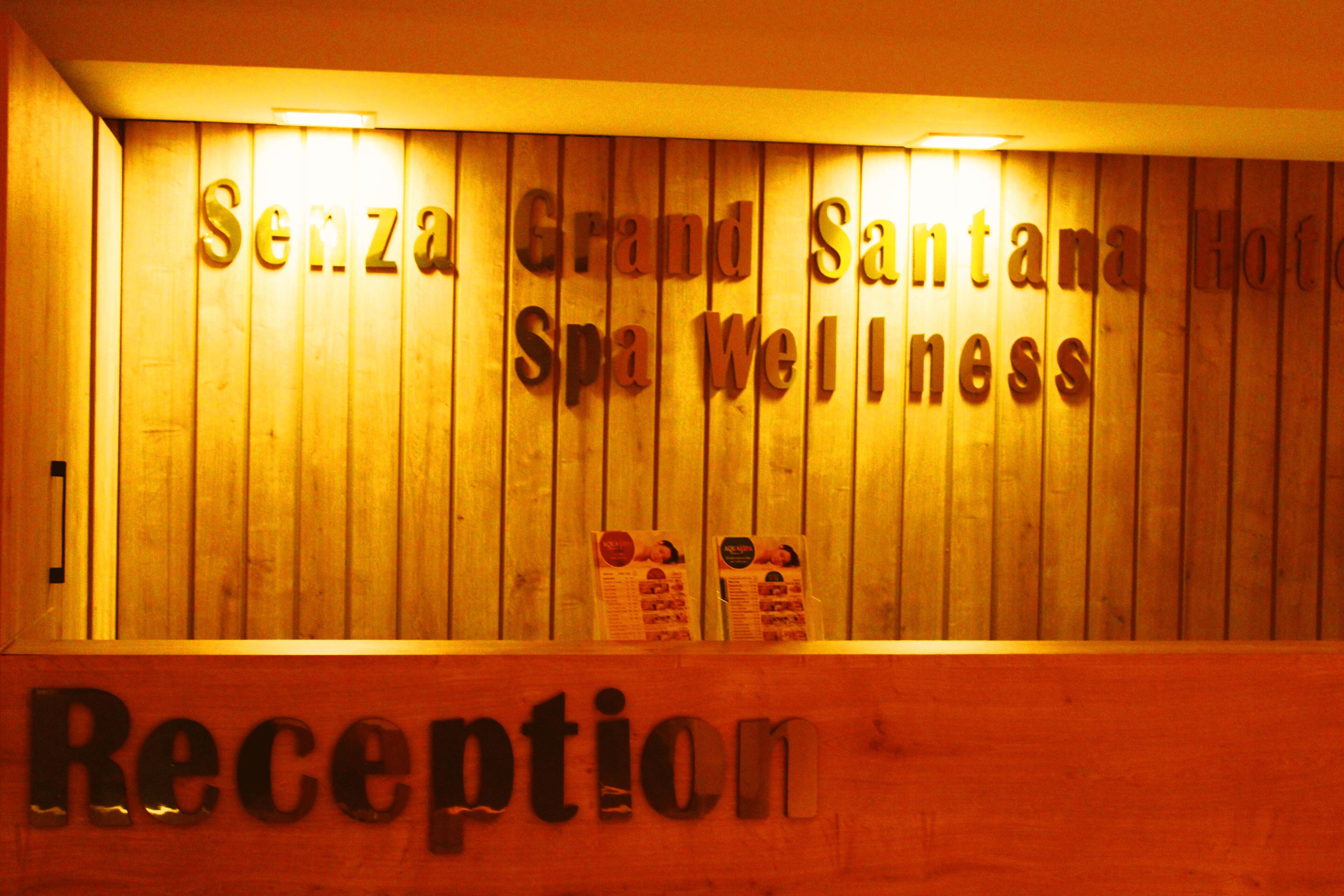 Uma área de recepção com parede de madeira exibindo o nome 'Senza Grand Santana Hotel Spa Wellness'.