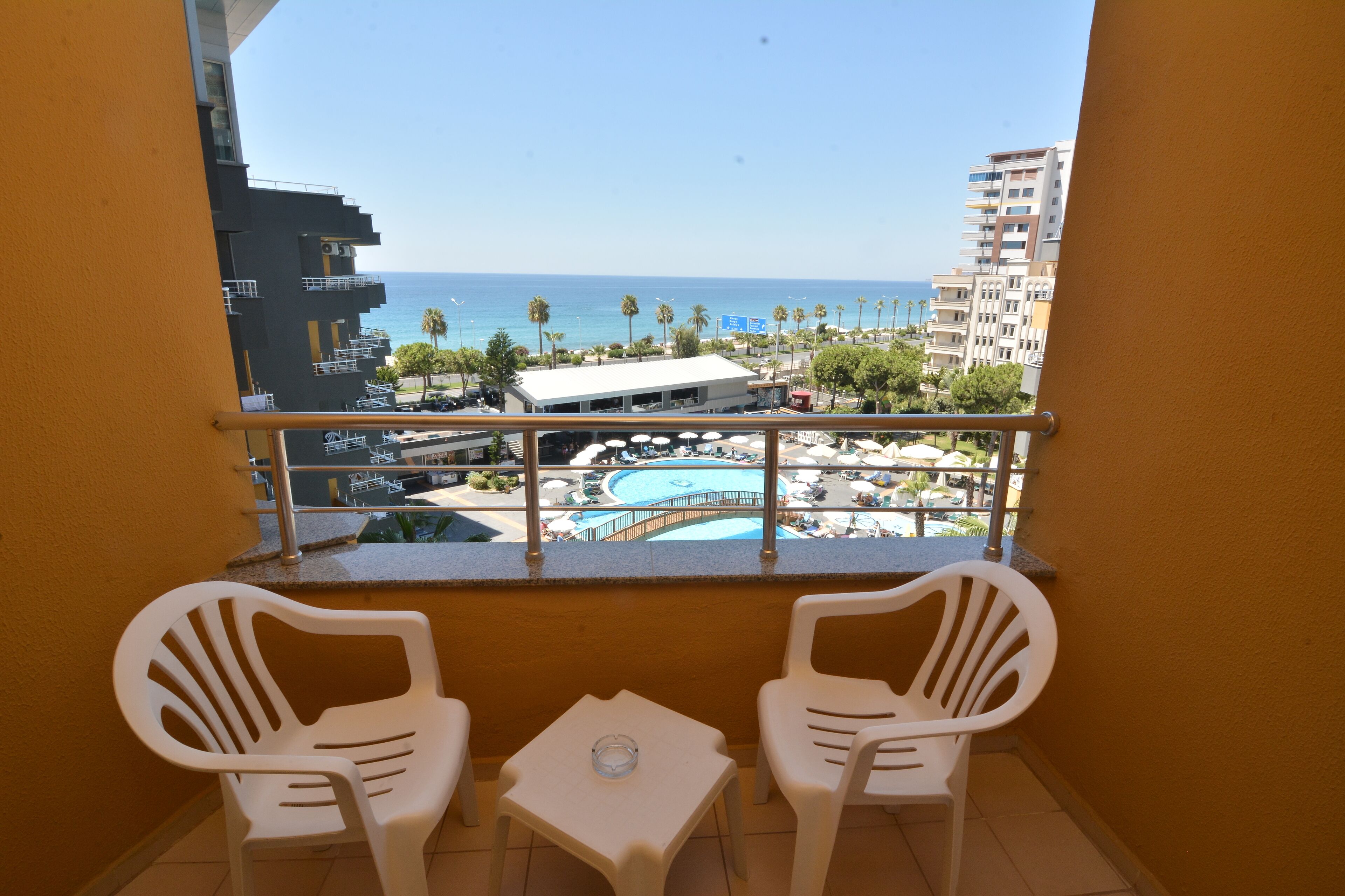 A balcony with two white plastic chairs and a small table, overlooking a pool area with a view of the ocean and a building in the background.