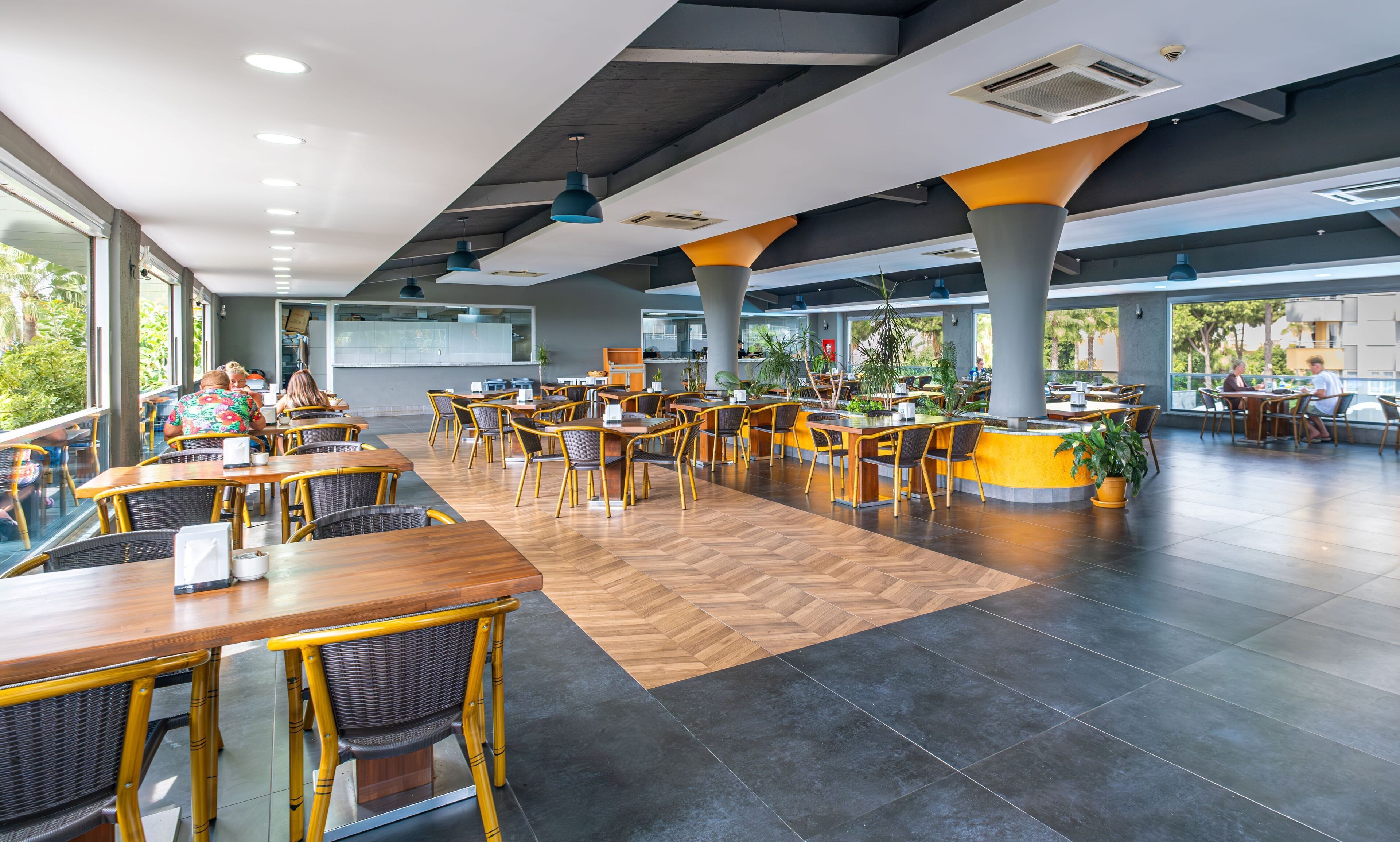 A spacious indoor dining area with wooden tables and chairs, large windows, and potted plants.