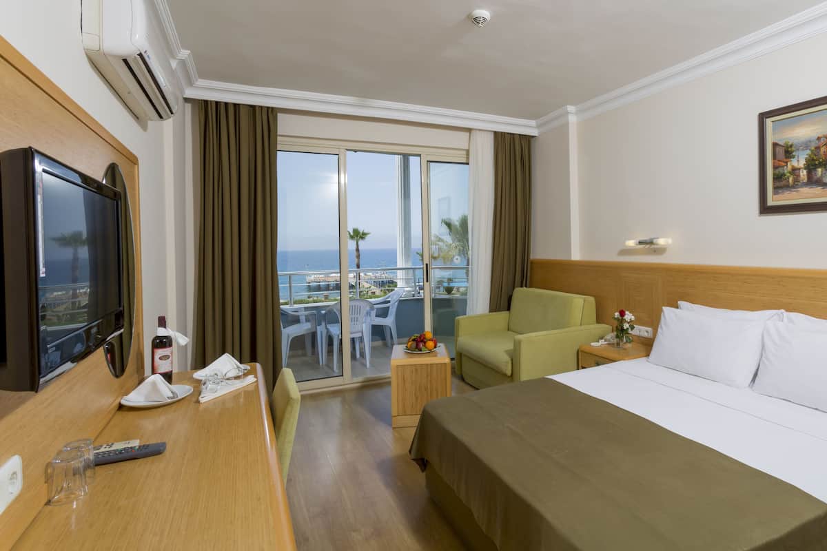 Une chambre d’hôtel avec un lit, une télévision, un bureau avec une télécommande, un verre d’eau, un vase avec des fleurs, une chaise, une petite table et une vue sur la mer depuis un balcon.