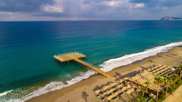 Vista aérea de una playa con un muelle de madera que se adentra en el mar, bordeado de tumbonas y sombrillas.
