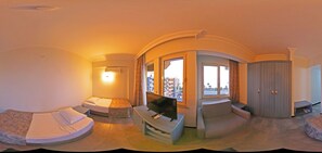 Una vista panorámica de una habitación de hotel con distribución curva, una cama, zona de estar, televisor y vistas de la ciudad a través de amplias ventanas.
