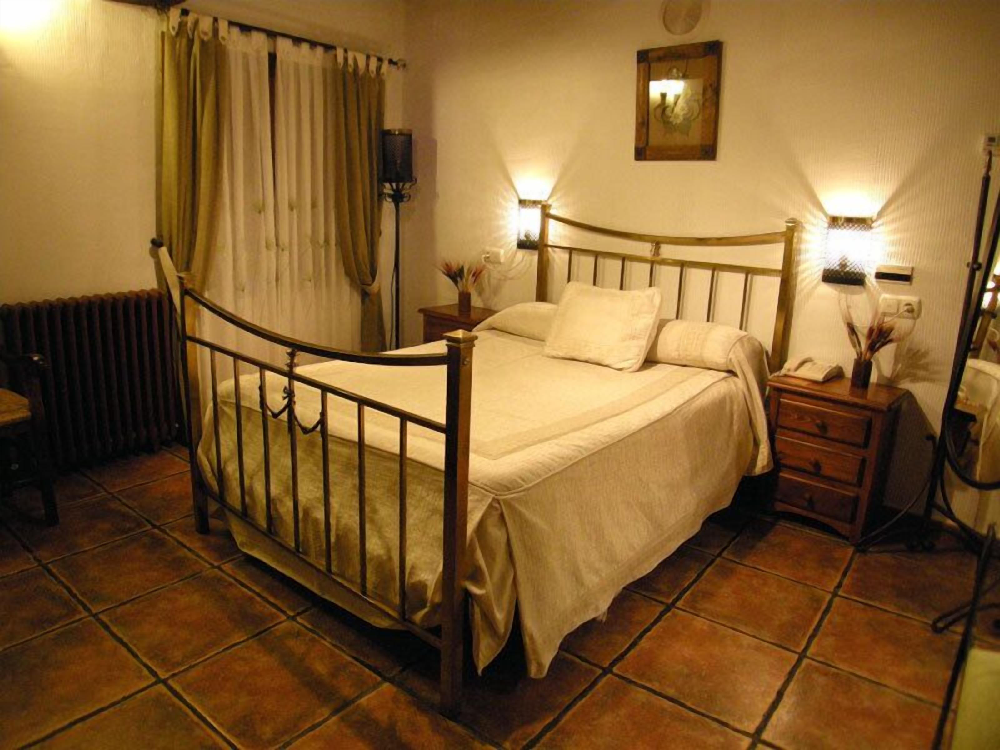 A bedroom with a wooden four-poster bed, a radiator, a nightstand with a lamp, and a wall-mounted clock.