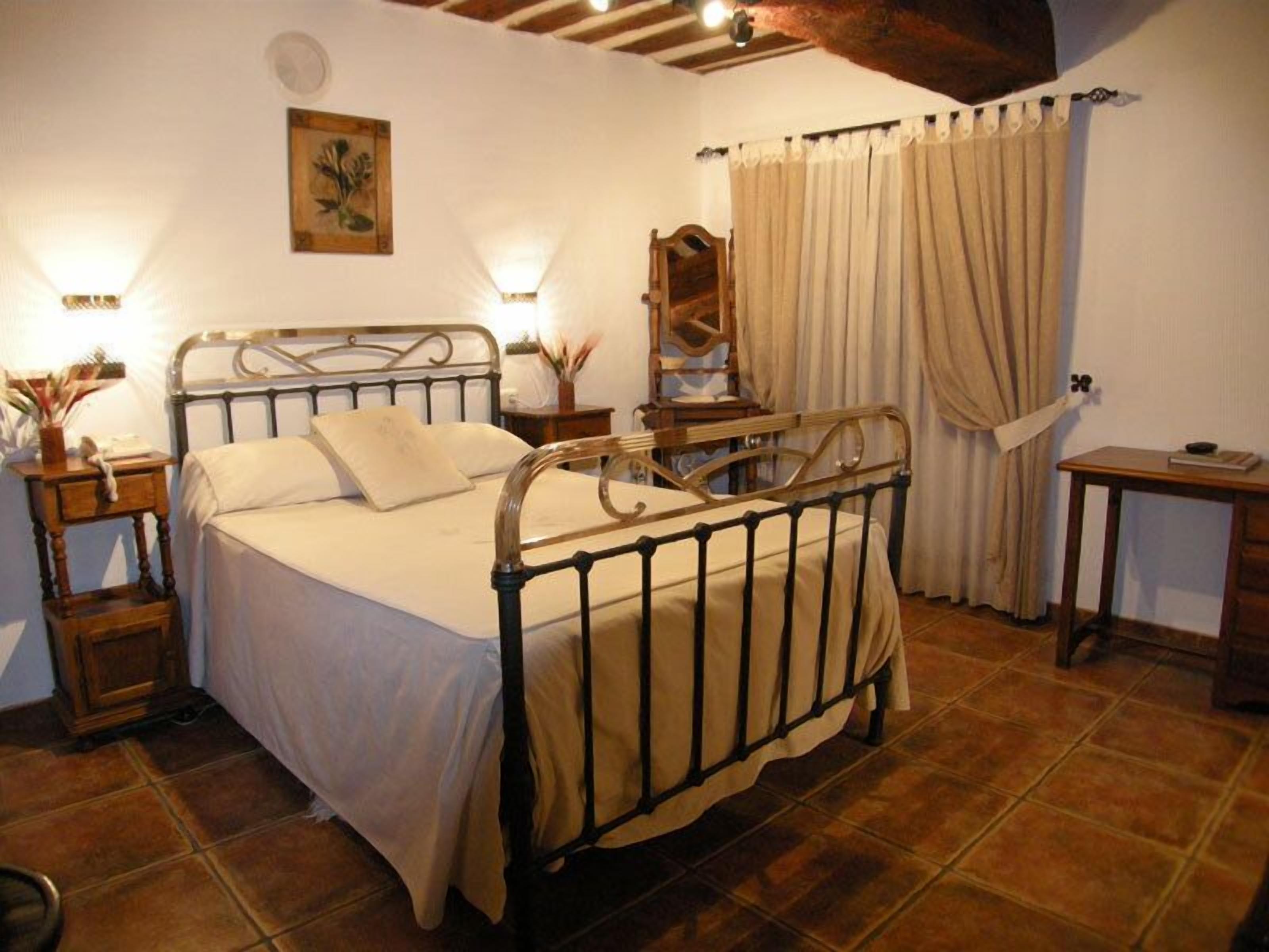A bedroom with a metal-framed bed, a bedside table with a lamp, a nightstand, a wooden chair, a mirror, and a window with curtains.