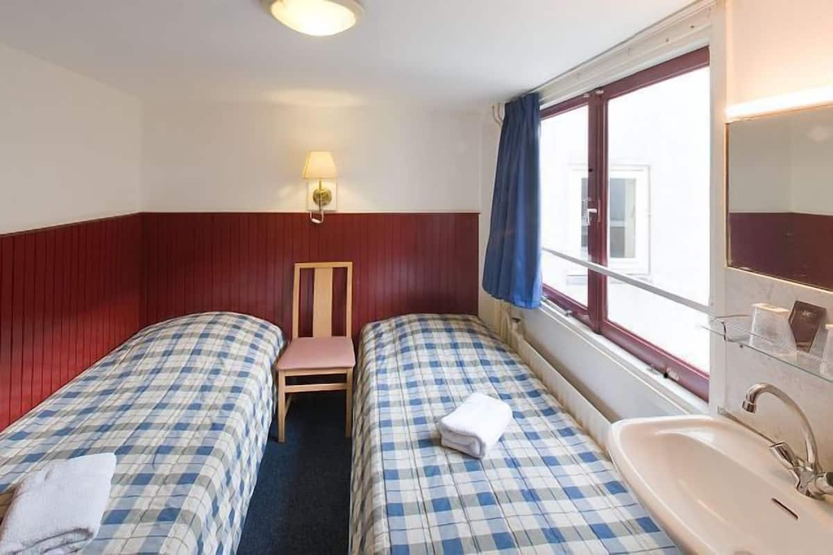 Habitación de hotel con dos camas, una ventana con cortinas, una lámpara de pared y un lavamanos.