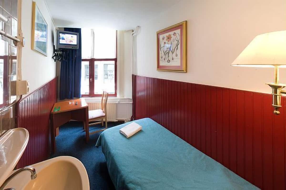 Una habitación de hotel pequeña con una cama individual, un escritorio con una silla, televisor, ventana con cortinas y baño con lavamanos y espejo.