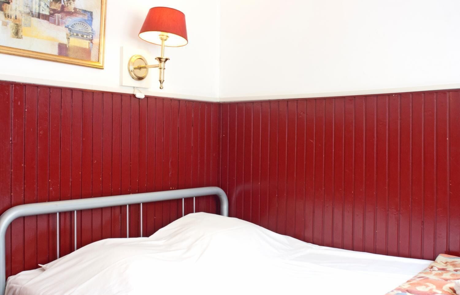 A room with a metal bed frame, a red wall, and a wall-mounted lamp.
