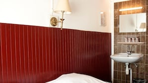 Habitación de hotel con pared revestida de paneles rojos, cama blanca, lámpara de pared y lavamanos con espejo, además de artículos de aseo en un estante.