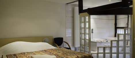 Quarto de hotel com cama, poltrona, janela com vista e banheiro visível por uma porta de vidro.