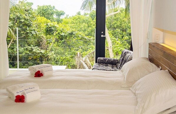 Une chambre à coucher avec un lit, des oreillers et une vue sur une végétation luxuriante, visible par la fenêtre.