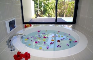 Een moderne badkamer met een groot, ovaal bad vol water en drijvende bloemen, een chromen kraan en uitzicht op weelderige begroeiing door grote ramen.