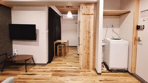 A hallway with wooden flooring, a television mounted on the wall, a small table with a chair, a washing machine, and a wooden door.