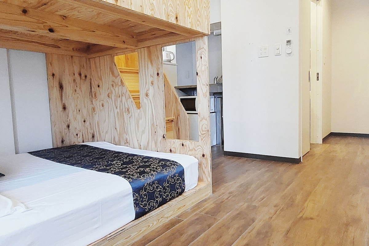 A modern room with a wooden bunk bed, a white wardrobe, and a kitchen area in the background.