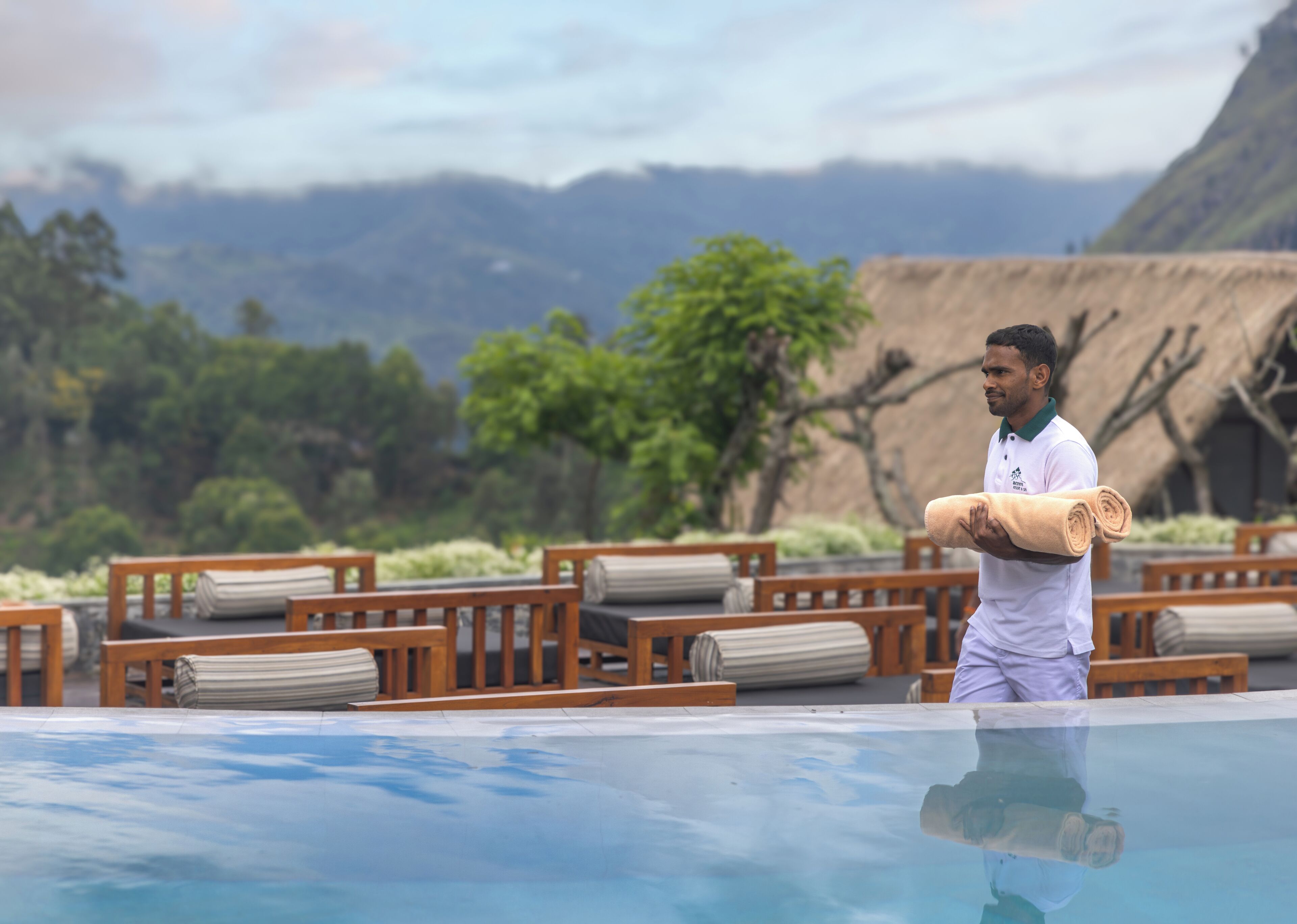 A person standing by a pool with wooden lounge chairs and a thatched-roof structure in the background.