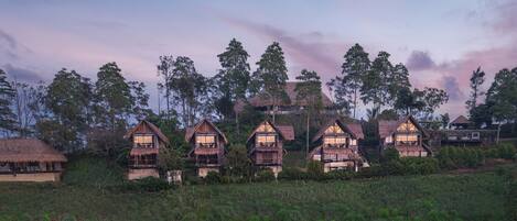 A row of thatched-roof cottages with balconies, set in a grassy field with trees in the background.