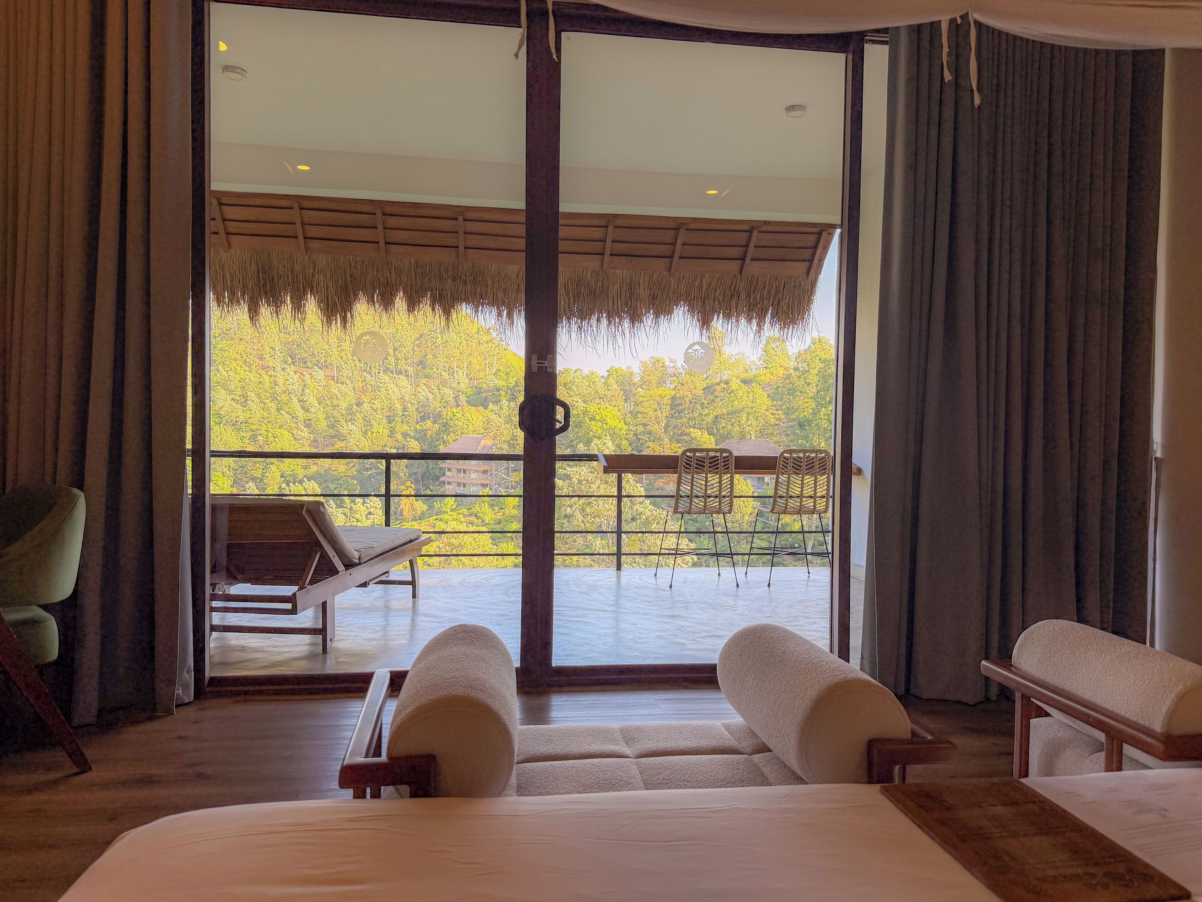 A room with a bed, two chairs, a table, and a balcony with a view of a forest.