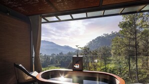 A modern bathroom with a wooden bathtub, a chair, and a large window offering a view of a mountainous landscape. - 98 Acres Resort & Spa (Ella)