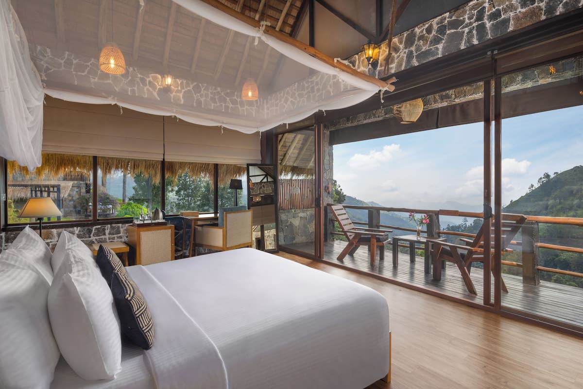 A spacious bedroom with a large bed, a desk, and a balcony with a view of the mountains.