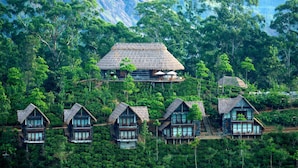 A cluster of thatched-roof cottages surrounded by dense forest, with a prominent house featuring a balcony and a staircase leading to a higher level. - 98 Acres Resort & Spa (Ella)