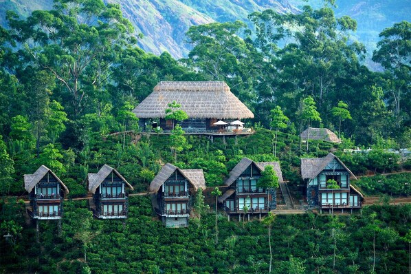 Un grupo de casas de campo con techo de paja rodeadas de un bosque denso, con una casa destacada que tiene un balcón y una escalera que conduce a un nivel superior.