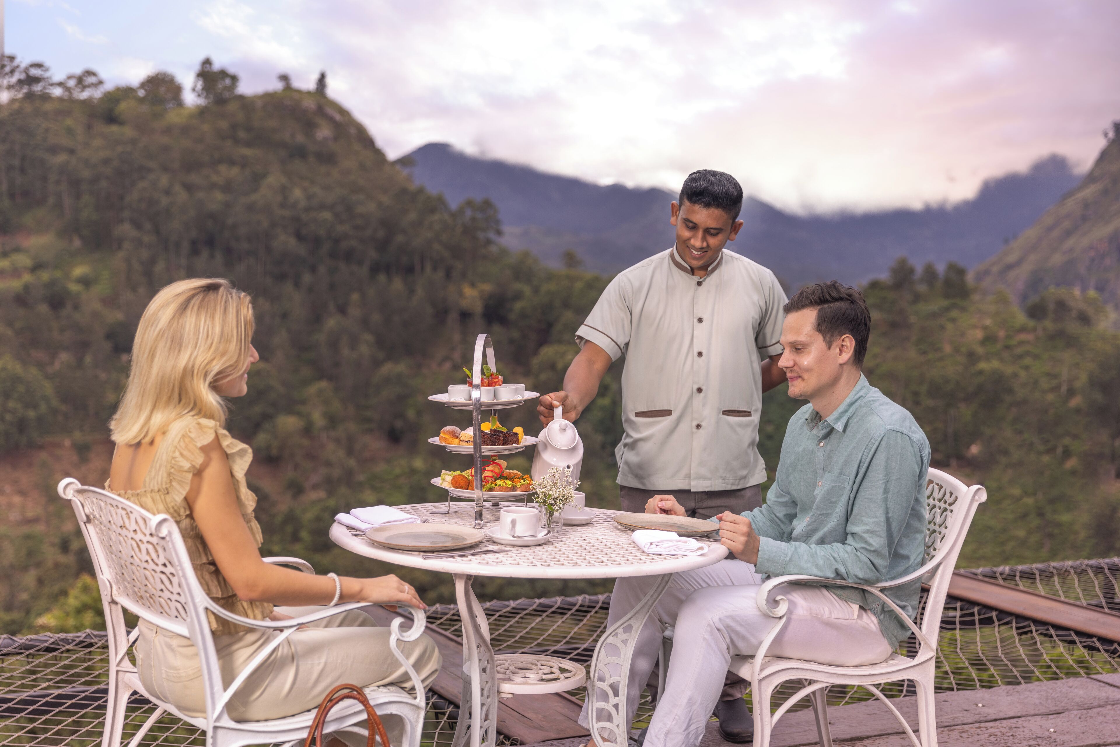 A table set outdoors with a scenic mountain backdrop, featuring a tiered dessert stand and a teapot.