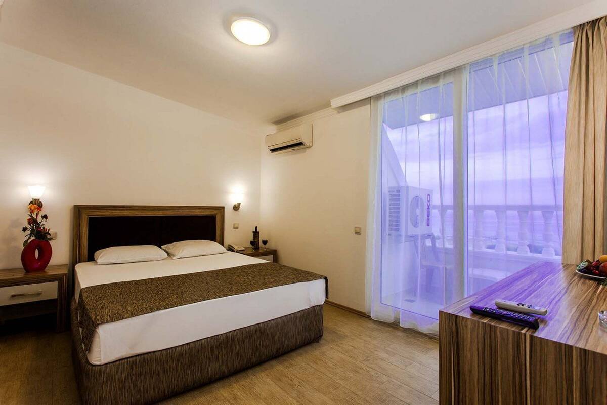 A hotel room with a large bed, a bedside table, a nightstand, a lamp, a vase with flowers, a window with sheer curtains, and a view of buildings outside.